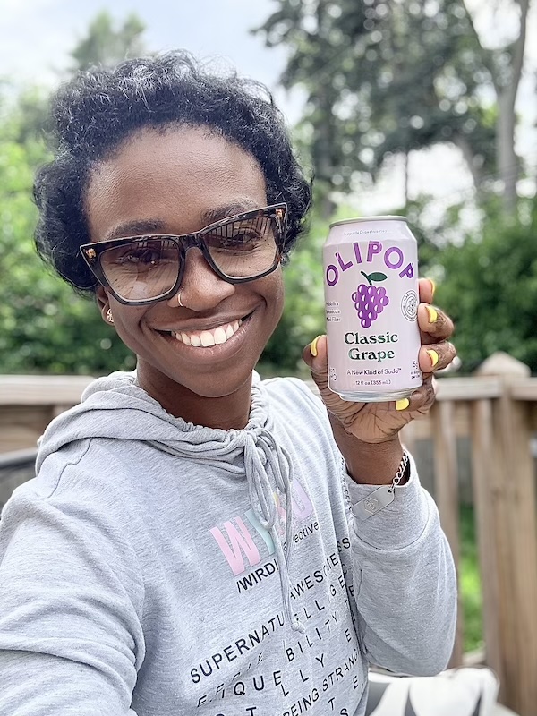 Ceva smiling and holding an OLIPOP Classic Grape can outdoors
